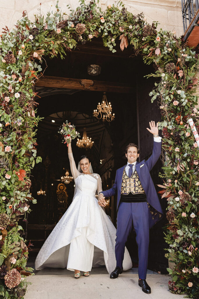 Novios saliendo de edificio con ramo en alto y arco floral granatum